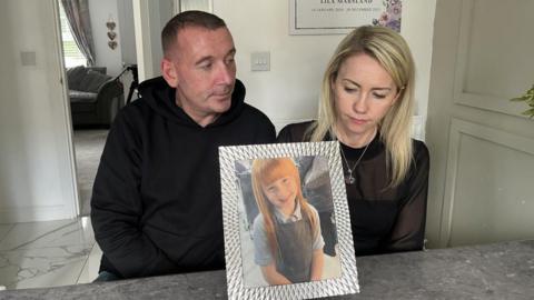 Lila's parents are looking at photograph of Lila in a silver frame. They are both sitting on stools in their kitchen. 