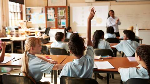 The image shows the rear view of girl raising her right hand while sitting next to friend during a lecture in a full classroom.