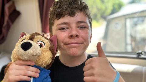 Leo O'Brien smiles at the camera and gives a thumbs up as he holds up a teddy bear. He is wearing a black Harry Potter t-shirt.