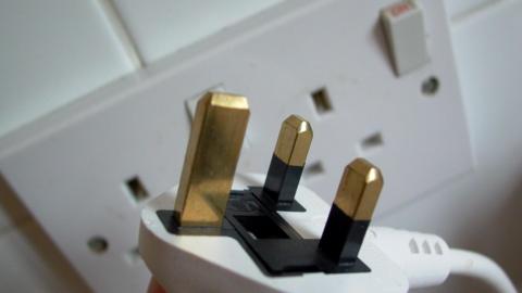 A close-up of a white three-point plug in front of a white wall socket.