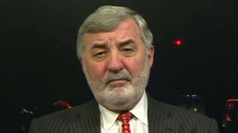 Lord Alderdice has grey hair parted to the side with a grey beard and is wearing a pin striped dark suit