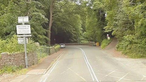 The road at the MER crossing - it bend off to the left and there are green trees on both sides and a sigh that says Welcome to Maughold on the left.