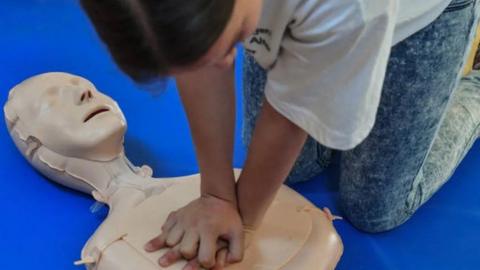 A rubber CPR dummy on a bright blue floor with a person's hands clasped in the centre of the dummy's chest practicing CPR.