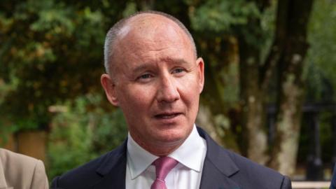 Jim Gavin, a man with receding, short, grey hair, addressing reporters outside a hotel in County Cork last week.  He is wearing a navy suit, a white shirt and a pink tie.  There are tall, mature trees in the background. 