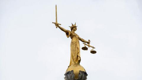 Gold Lady of Justice statue with a sword in one hand and scales in the other. She is wearing a crown.