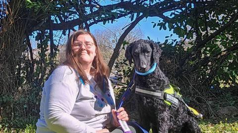 Amy Harris with her dog Wish. Amy has brown hair and is wearing glasses. Her dog has black fur and a harness.