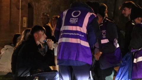 A group of volunteers in purple reflective jackets, with Street Angels Chester logos in white lettering, speak to a group of younger people sitting on a bench on a street at night time. A young man is drinking from a bottle of water.