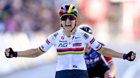 Kim Le Court, wearing a largely white cycling jersey, multicoloured helmet and sunglasses, sits up on her bike and puts her arms out wide in celebration after winning a cycling race