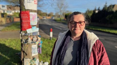 Keena Entwistle, a woman in a red coat, blue scarf and grey top. She is stood beside a road and a tree with flowers and signs attached to it. She is looking at the camera.
