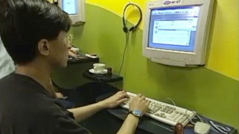 A man with dark hair, glasses and black top, sits at a computer terminal in a cafe, with his hands on the keyboard, a monitor ahead mounted on the wall and a pair of headphones hanging on the wall on a hook.  To his left sits a coffee cup on a saucer.