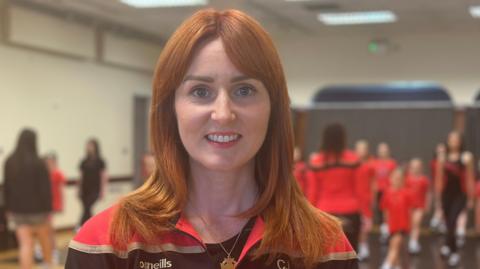 Catherine Lamont has shoulder length auburn hair and is wearing a tracksuit which is black with red shoulders and gold trim. In the blurred background girls can be seen practicing Irish dancing, also wearing red and black