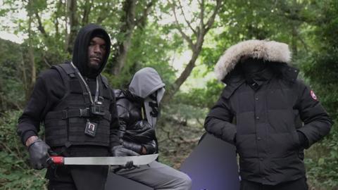 Faron Paul who runs his own knife amnesty company holds a large knife. He is standing in a woodland area next to two men whose faces are covered. The man on the right is "Matthew" (not his real name) who has handed in the knife.