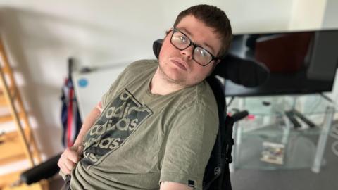 George Grimshaw in a wheelchair in his flat. There is a television on a glass stand behind him.
