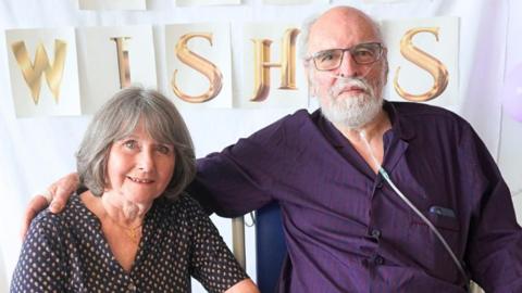 A woman with shoulder-length grey hair in a spotted dress sitting down next to a bald elderly man with a white beard who is wearing a purple and blue shirt and has a nose tube in.