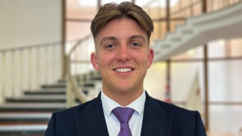 A man with parted fair hair, cropped at the sides, smiles at the camera in front of a curved staircase inside a council building. He is wearing a dark jacket, white shirt and purple tie.