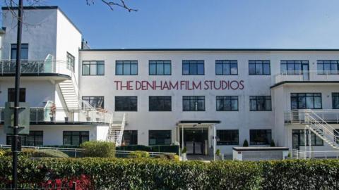 A former film studio, a white building with writing in it in red. It has black windows, a main entrance and is in the Art Deco style. It has a front door and steps either side. There are bushes outside the front of the building. 