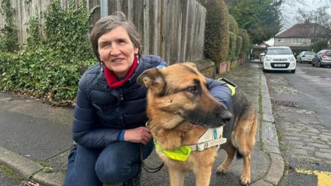 A woman with short grey hair wearing a navy jeans with a navy coat and red scarf crouched down on the pavement beside her guide dog, a German Shepherd wearing a hi-vis harness, who is looking to the right. Cars parked on the road in the background.