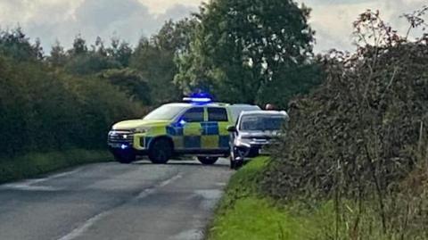 A police car and a truck parked down a country lane near Cirencester in Gloucestershire