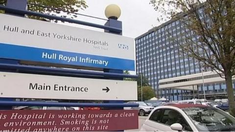 A blue and white sign directing visitors to the main entrance of Hull Royal Infirmary. The tall, glass-fronted hospital building is in the background. Cars are parked nearby.