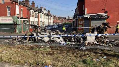 Police tape surrounds what appears to be an extinguished bonfire. It is possibly the burnt remains of a bus. A street of redbrick terraced homes is in the background.