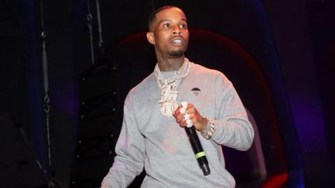 Rapper Tory Lanez is seen on stage holding a microphone. He is wearing a gray sweatshirt and multiple diamond necklaces and a big diamond ring on each finger. 