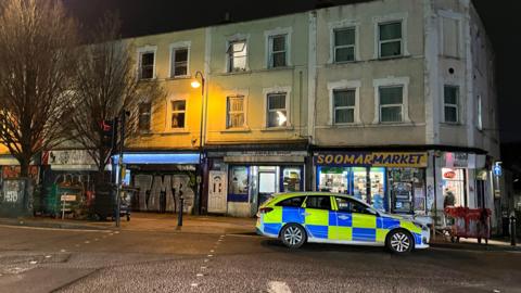 A police car parked on a street after dark with the road lit by a lamp-post. Shop fronts can be seen on the ground floor of a row of buildings with windows on the floors above and trees to the left.