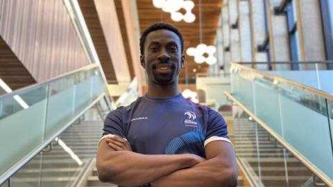 A man wearing a blue sports top crossing his arms, stood on a staircase