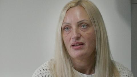 Jenni Smith with long blond hair wearing a white knitted crew neck jumper. She is sitting in an office with a white wall behind her. She is looking impassive.