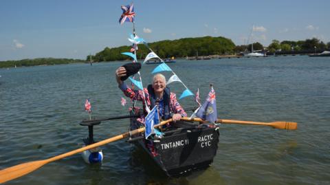 An elderly man in a home-made boat made from corrugated metal. He is waving his dark blue cap while wearing a Union Jack jacket, with Union Jacks on the boat.