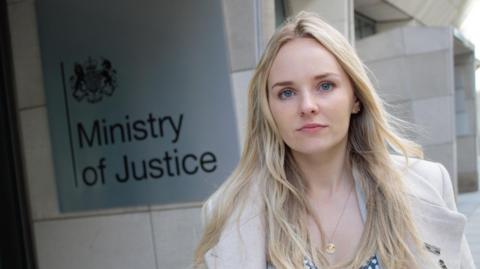 Harper has long blonde hair and blue eyes. She wears a white coat and stands next to the sign for the Ministry of Justice.