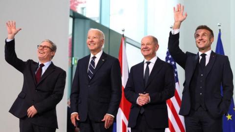 UK Prime Minister Keir Starmer,  US President Joe Biden, German Chancellor Olaf Scholz and French President Emmanuel Macron pose for a family photo session at the Chancellery in Berlin, 18 October. Starmer and Macron are holding their right hands in the air and waving. All four are smiling and wearing dark suits, and there are flags of the UK, US and EU behind them.