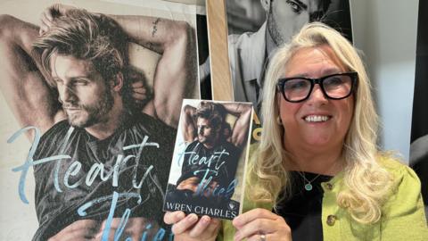 A woman with long blond hair is wearing black rimmed glasses a black top and lime green cardigan. She is smiling and holding a paperback book called Heart Thief in front of a large poster which has the book's front cover of a muscular man