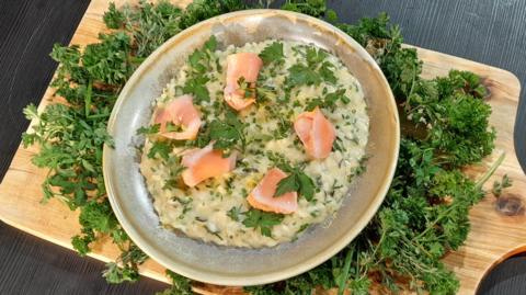 A plate of herby risotto, sitting on a bed of green herbs on a wooden board, with flaked pieces of salmon placed on top with green garnish.