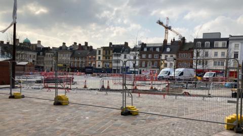 A wide shot of the market square undergoing construction with cobbled flooring, metal gates, cones, vans and cranes on show