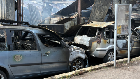 In the foreground, there are two severely fire-damaged vehicles parked beside a kerb: A grey MPV on the left has extensive fire damage, with burned paintwork, missing windows and a heavily charred roof.