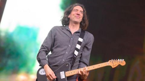 Gary Lightbody of Snow Patrol performs during BBC Radio 2 Live in the Park at Moor Park, Preston. He is pictured looking at the crowd while playing the guitar. He wears a dark grey shirt and has shoulder length brown hair.