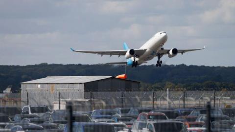 A large white passenger plane with a light blue tail taking off from an airfield. It can be seen above a large grey shed, a barbed wire fence and dozens of parked cars.