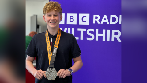 Roman, a teenager with ginger hair, standing in front of a purple BBC Radio Wiltshire sign, smiling wide and bright, and wearing his silver medal around his neck while holding it on each side.
