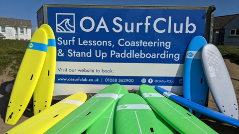 A blue and white trailer for OS Surf Club with yellow, green and blue surfboards in front of it.