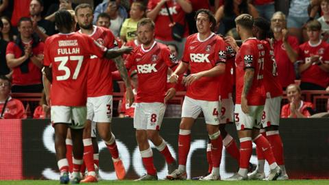 Seven players from Charlton Athletic smile and celebrate with each other after a goal.