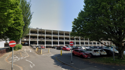 An image of a grey multi-storey car park with green trees either side