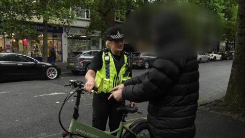 A female police officer called Sgt Clare Moore holds an illegal e-bike on a Plymouth street during a police operation. She is talking to a person - possibly the bike's rider - who is wearing a black coat with its hood covering his head. His head has been blurred. Sgt Moore has a yellow high-vis police jacket on. The e-bike is green and has emovement written on it in white letters. Its wheels and handlebars are black.