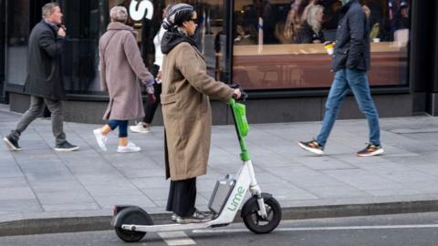 A woman rides a Lime electric scooter on a city street in front of a Starbucks, while pedestrians walk past and customers sit inside the café.