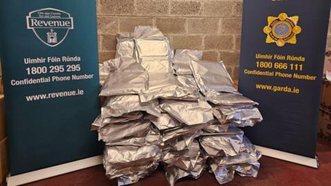 Two large posters are next to a large seizure of drugs which is in silver packets. The posters are dark blue and turquoise with 'revenue customs service' and 'An Garda Síochána' written on it with their logos.