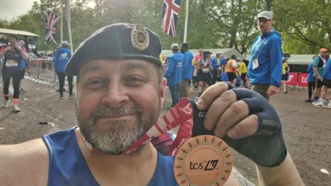 Mike Ryan wearing his beret while holding up the medal around his neck. Other competitors and spectators are in the background.