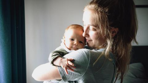 A young mum with light brown hair and wearing a grey T-shirt holds her baby in her arms and looks at him with her head turned to one side, standing in front of a window with daylight streaming through.