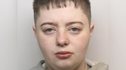 A mugshot of a pale person with short hair, which is buzzed at the side and slightly on the forehead from above. Her eyes are slightly narrowed and she has a neutral or tired expression. She has a tattoo on the side of her neck and a grey hoodie on.