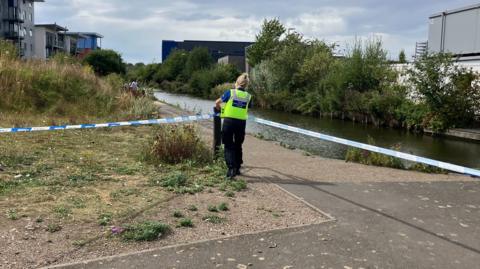 A woman in a high visibility vest attaches tape to a post with a canal and towpath visible and shrubbery as well as buildings in the distance