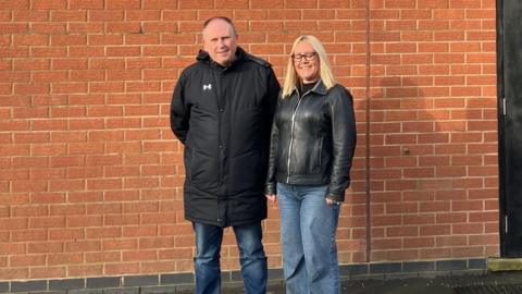 A man in a long black coat and jeans and a woman with long blonde hair, black leather jacket and jeans stand in front of the brick wall of a sports club building