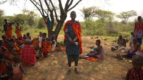 Women in traditional clothing are sitting in a clearing under a tree. They are holding medicine.